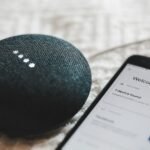 A charcoal Google Home Mini smart speaker next to a smartphone showing a smart home app