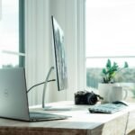Laptop, monitor, webcam, and plant on a wooden desk in a sunlit home office, illustrating home office gadgets for remote workers