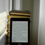 Kindle e-reader sitting on a windowsill next to a stack of books, illustrating e-readers compared for reading